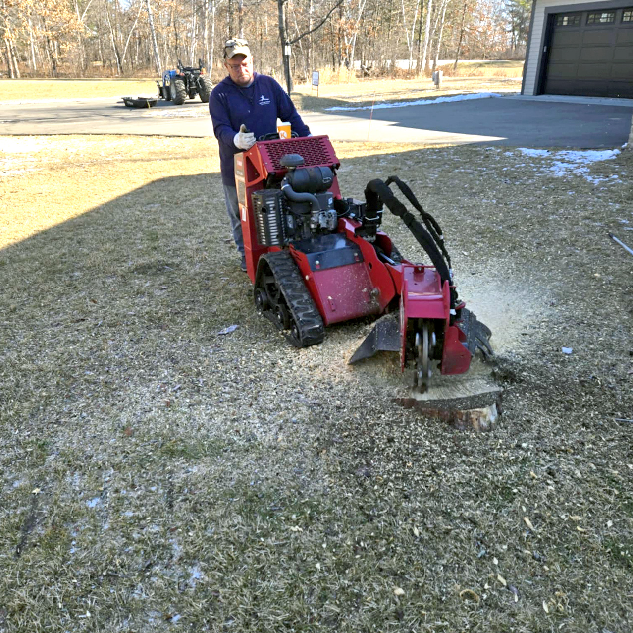 A person operates a red stump grinder machine on a grassy yard, grinding down a tree stump with wood chips scattered around. A driveway and garage are visible in the background.