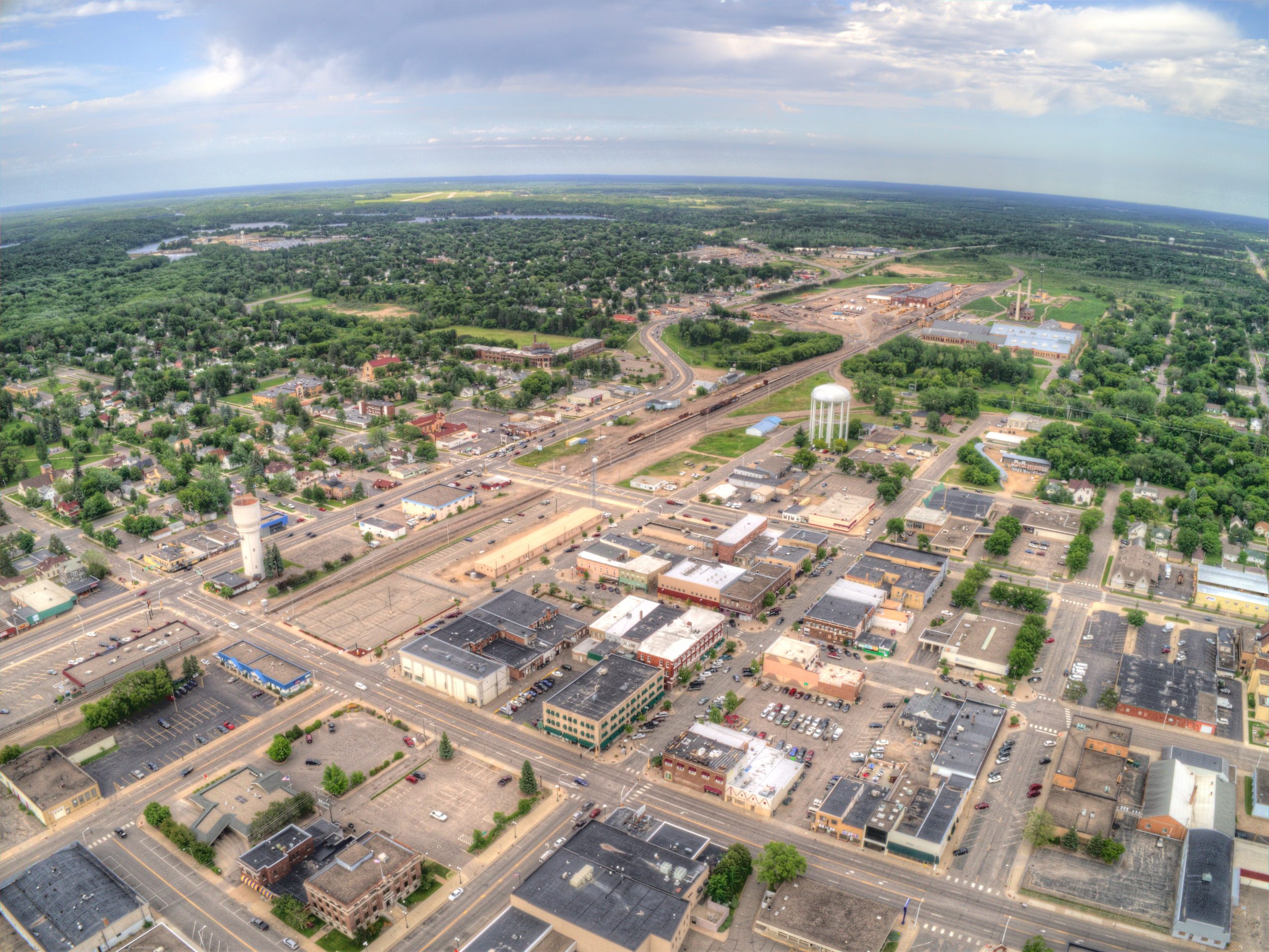 Aerial view of a small city with grid-patterned streets, commercial buildings, water towers, green trees, and open land stretching to the horizon under a partly cloudy sky.